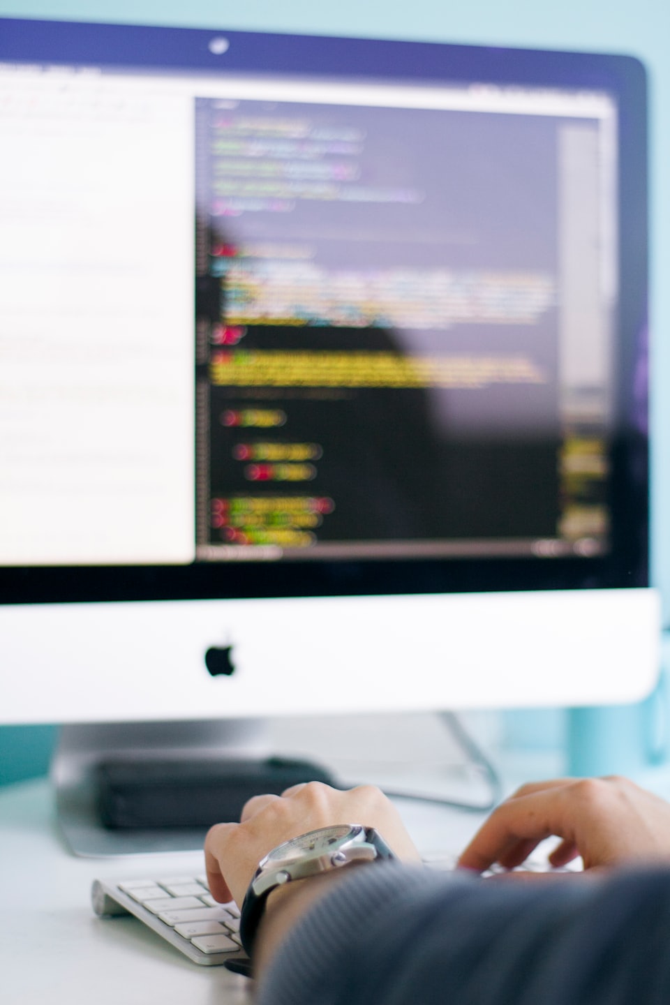 Hands typing on a white keyboard with a monitor displaying code.